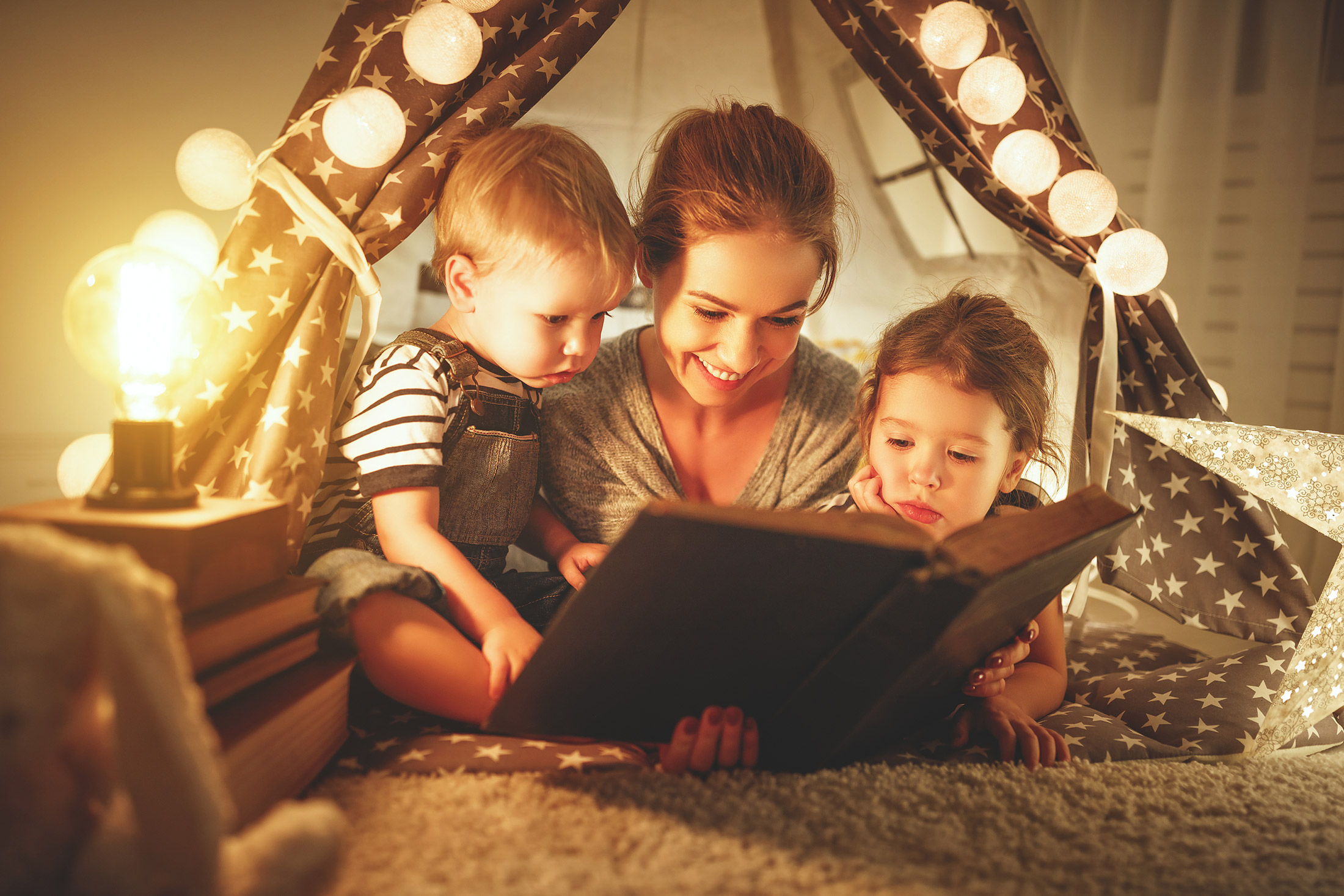 Eine Frau liest einem Jungen und einem Mädchen aus einem Buch vor. Sie bringt ihnen spielerisch das lesen bei.