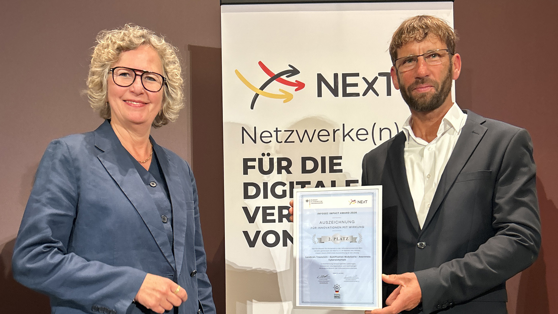 Bei der Verleihung des InfoSec Impact Award 2026 in Berlin: Astrid Schumacher, Fachbereichsleiterin im Bundesamt für Sicherheit in der Informationstechnik (BSI) und Vorständin von NExT e. V., übergab die Auszeichnung an Claus Hofmann, Informationssicherheitsbeauftragter und CISO des Landkreises Traunstein. Der Landkreis Traunstein erreichte mit seiner „Gamification Modulserie – Awareness Cybersicherheit“ den 2. Platz.